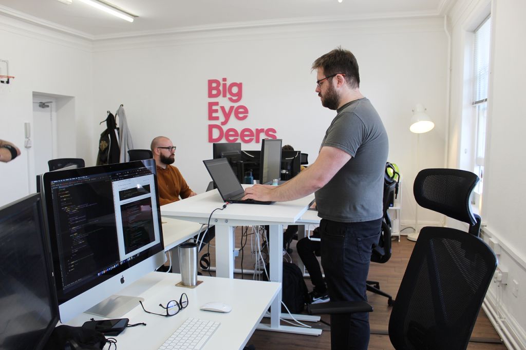 A side view of Pete, in the office on a computer with a pink sign behind saying 'Big Eye Deers'